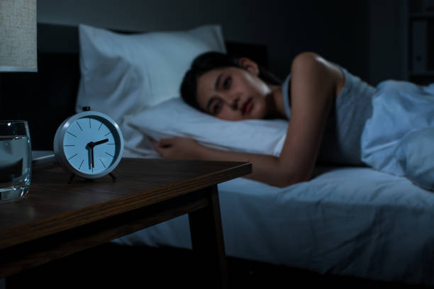Woman lying awake in bed at night next to a glass of water and a small alarm clock on a bedside table.