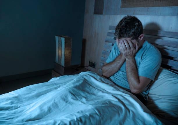 Man sitting up in bed at night, face buried in his hands, looking distressed/overwhelmed.